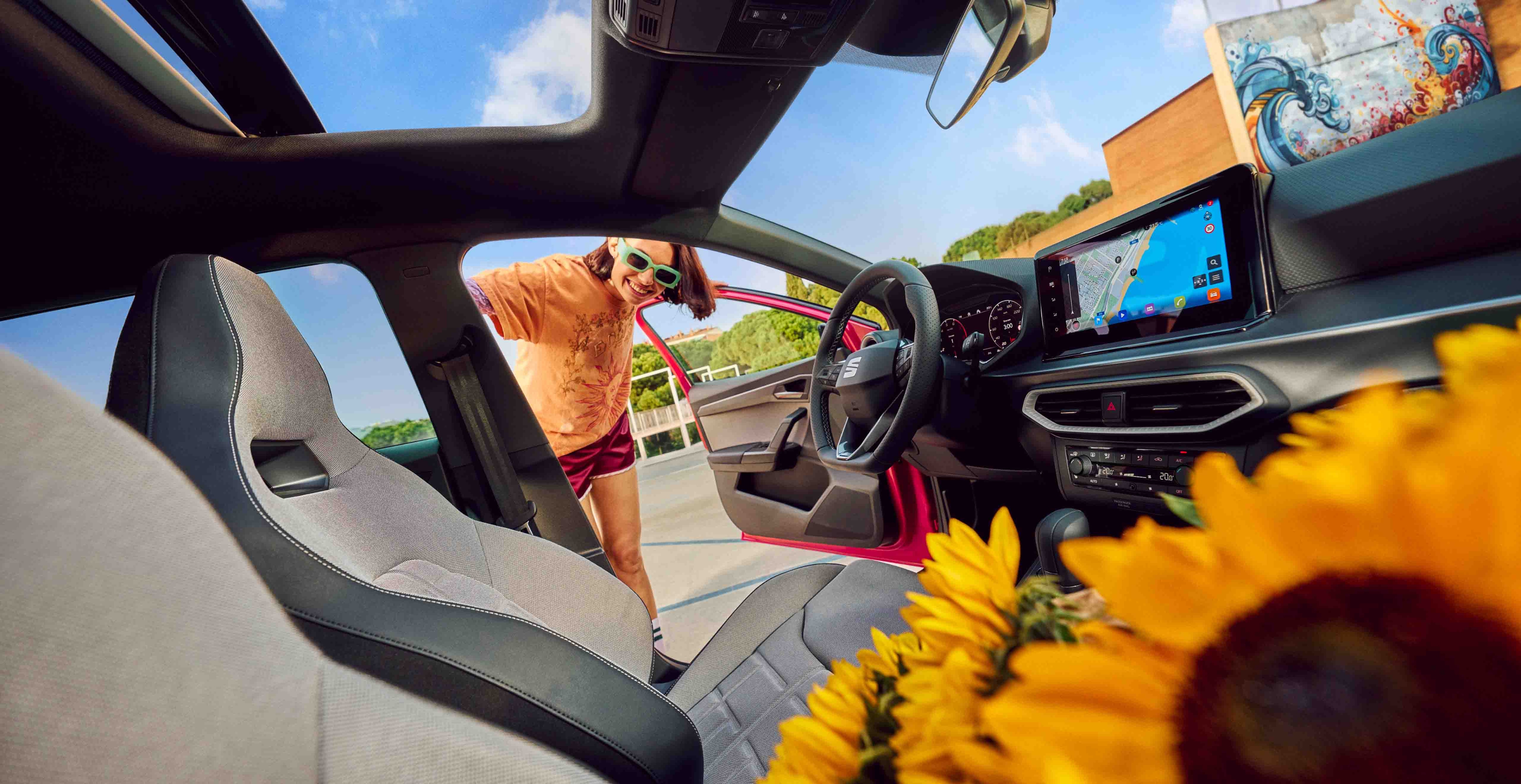 Vue de l’habitacle d’une SEAT avec toit panoramique et tableau de bord digital, une femme à la portière et des tournesols au premier plan.