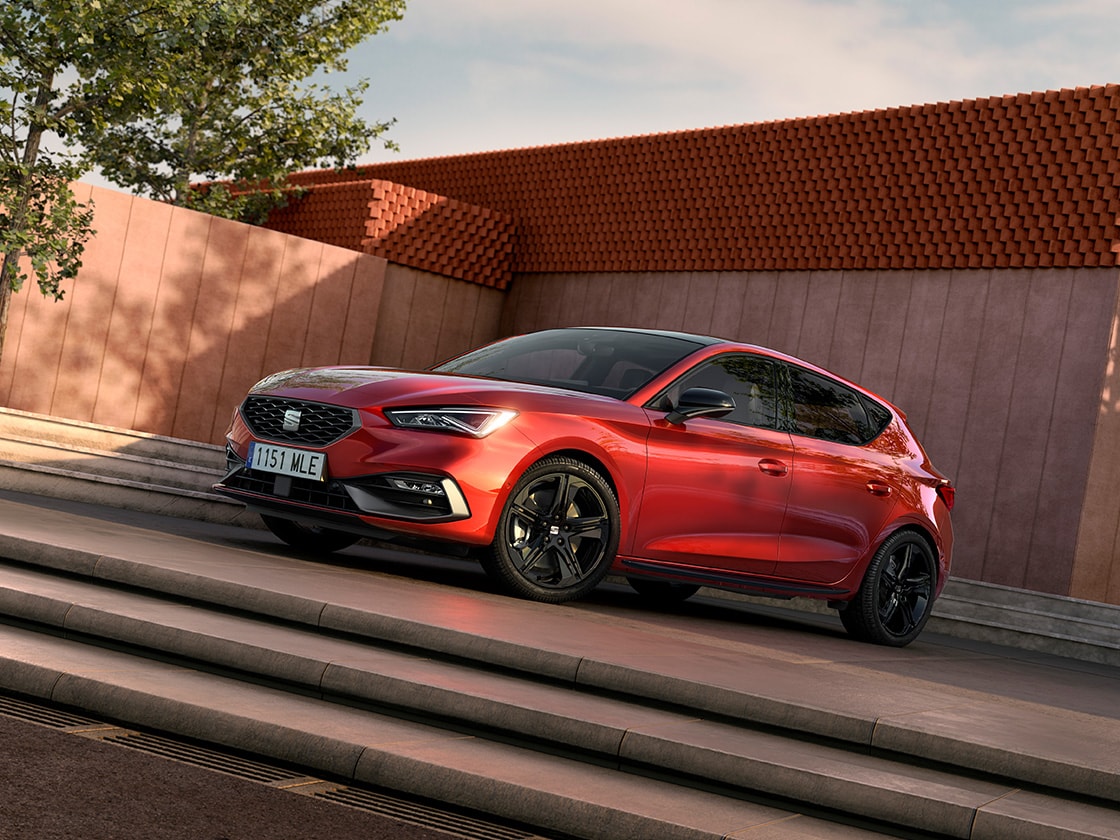 Vue d'une SEAT Leon NF 5D garée en extérieur entre des escaliers, avec un immeuble et un arbre en arrière-plan.