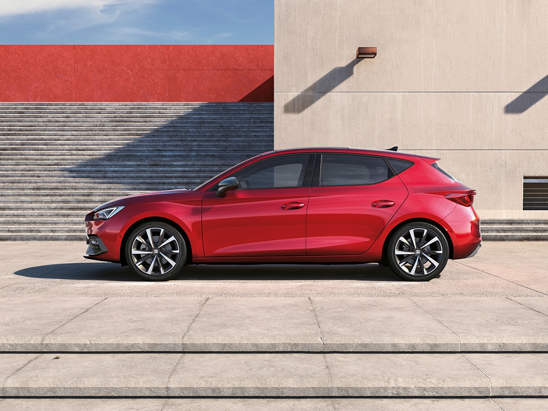 Vue latérale d'une SEAT Leon NF 5D en rouge désir garée en extérieur au soleil avec un immeuble et des escaliers en arrière plan.