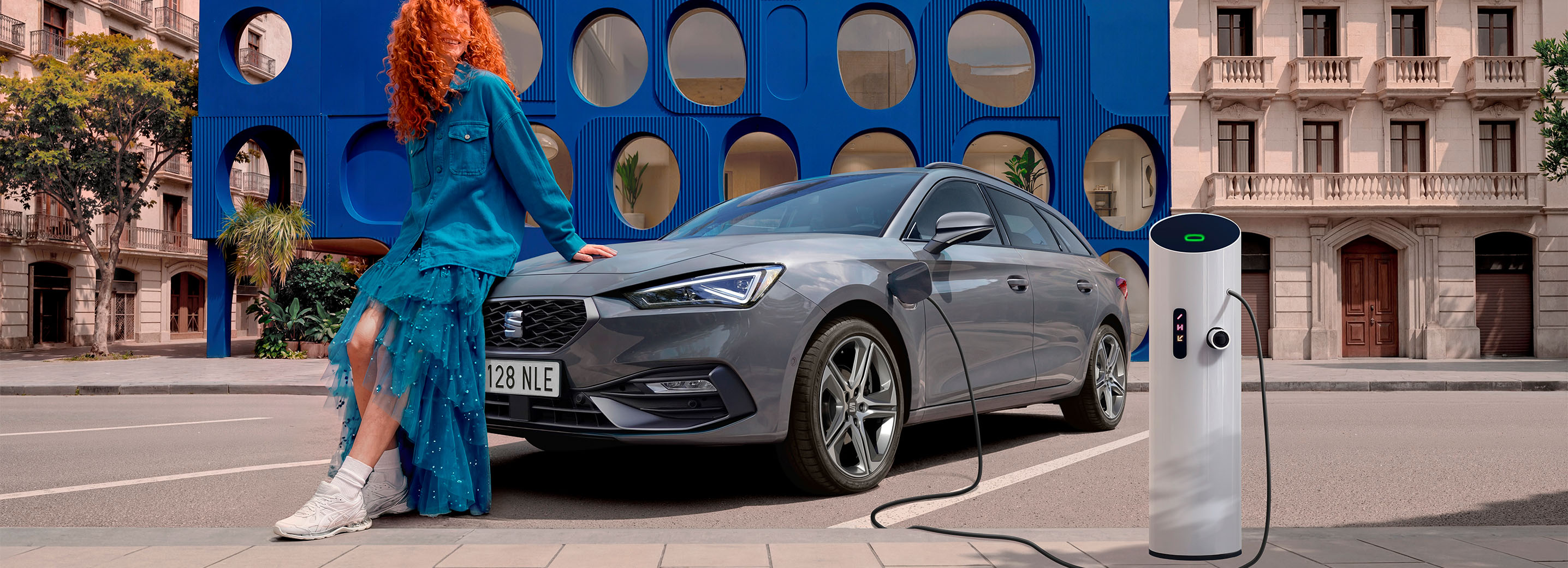 Vue de face 3/4 d'une SEAT Leon Sportstourer dans un environnement urbain avec des immeubles et une structure bleue en arrière-plan.