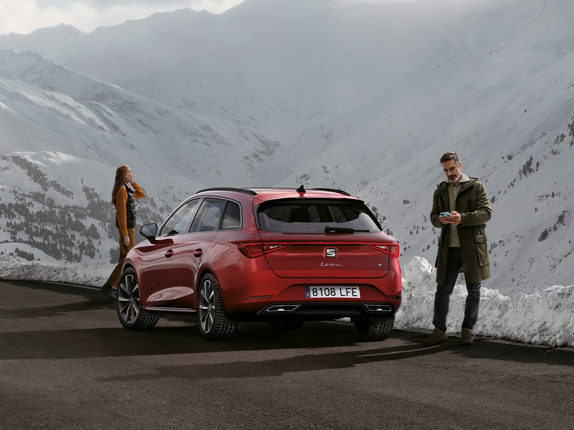 Une seat leon Sportstourer rouge garée sur une route de montagne, attendant les avantages du service de mobilité et de l’assistance routière seat. Deux personnes se tiennent à proximité.