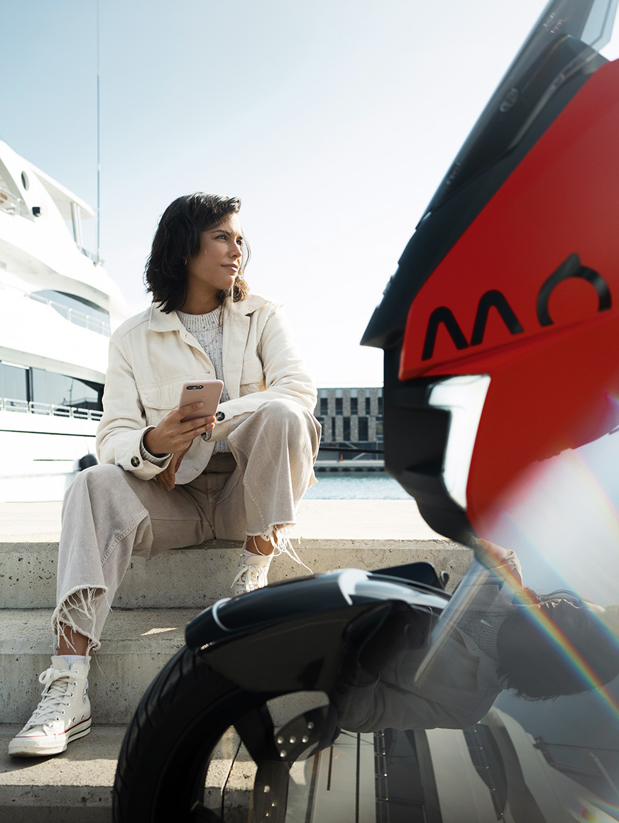 woman sitting by her parked SEAT MÓ 125 electric scooter at a harbour