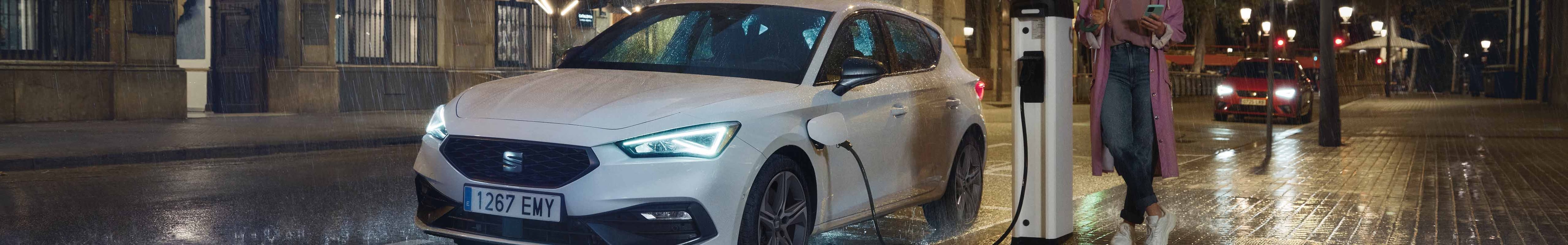 SEAT Leon en train de charger à une station de recharge publique dans une rue pluvieuse la nuit, mettant en évidence les avantages des véhicules hybrides.