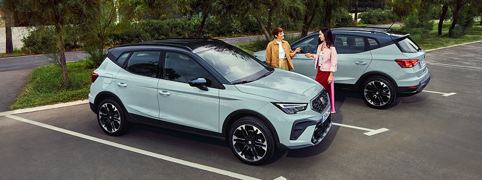 Vue de deux SUV compacts SEAT modernes avec une carrosserie bicolore, stationnés dans un parking en ville, avec en arrière-plan des gratte-ciel vitrés, des arbres verdoyants et un ciel bleu partiellement nuageux.
