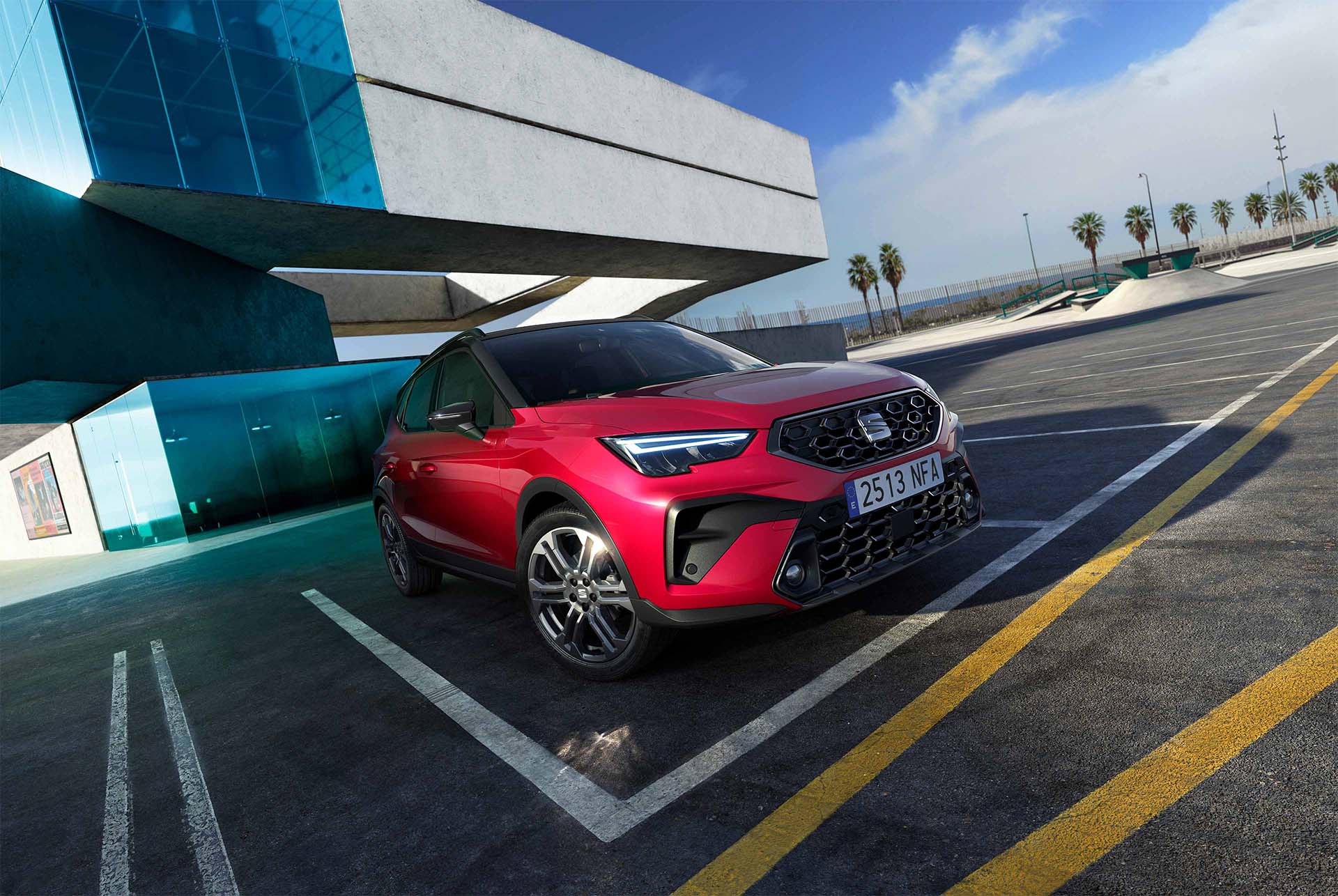 Vue en contre-plongée d’un crossover SUV urbain SEAT Arona rouge doté d’un toit noir, stationné sur une surface asphaltée sur laquelle sont dessinées des lignes jaunes. À l’arrière-plan apparaît un bâtiment moderne en béton blanc avec des panneaux de verr