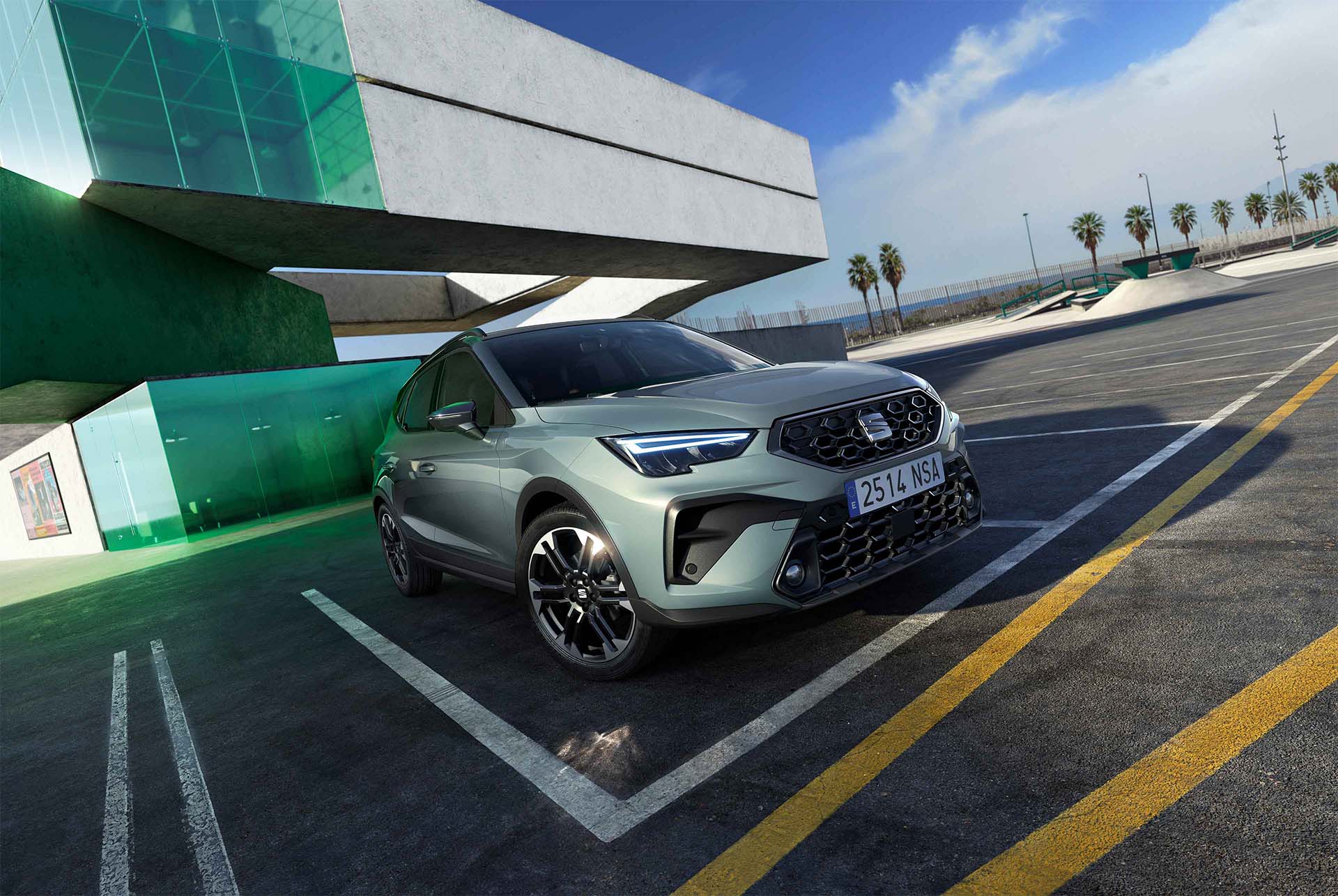 Vue en contre-plongée d’un crossover SUV urbain SEAT Arona gris, stationné sur une surface asphaltée sur laquelle sont dessinées des lignes jaunes. À l’arrière-plan apparaît un bâtiment moderne en béton blanc avec des panneaux dorés réfléchissants.