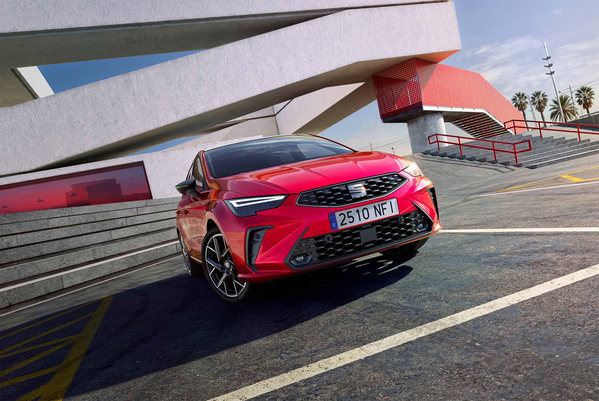 Vue d’une citadine SEAT Ibiza FR en finition rouge brillante, présentée dans un environnement urbain avec des structures angulaires en béton, sous un ciel bleu clair.