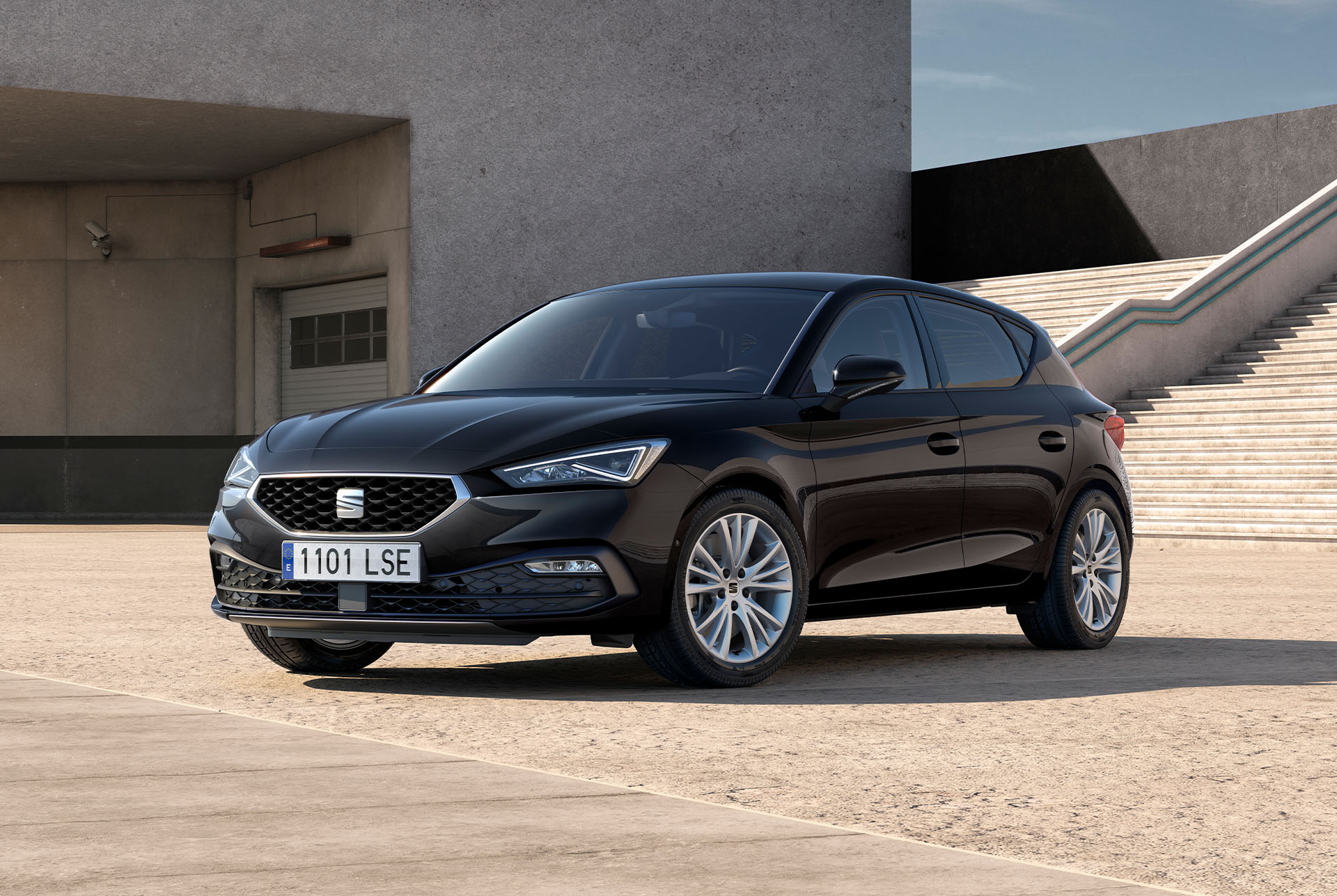 Une SEAT Leon 5 portes garée sur un parking, vue de trois quarts face devant un bâtiment en béton avec un grand escalier extérieur.