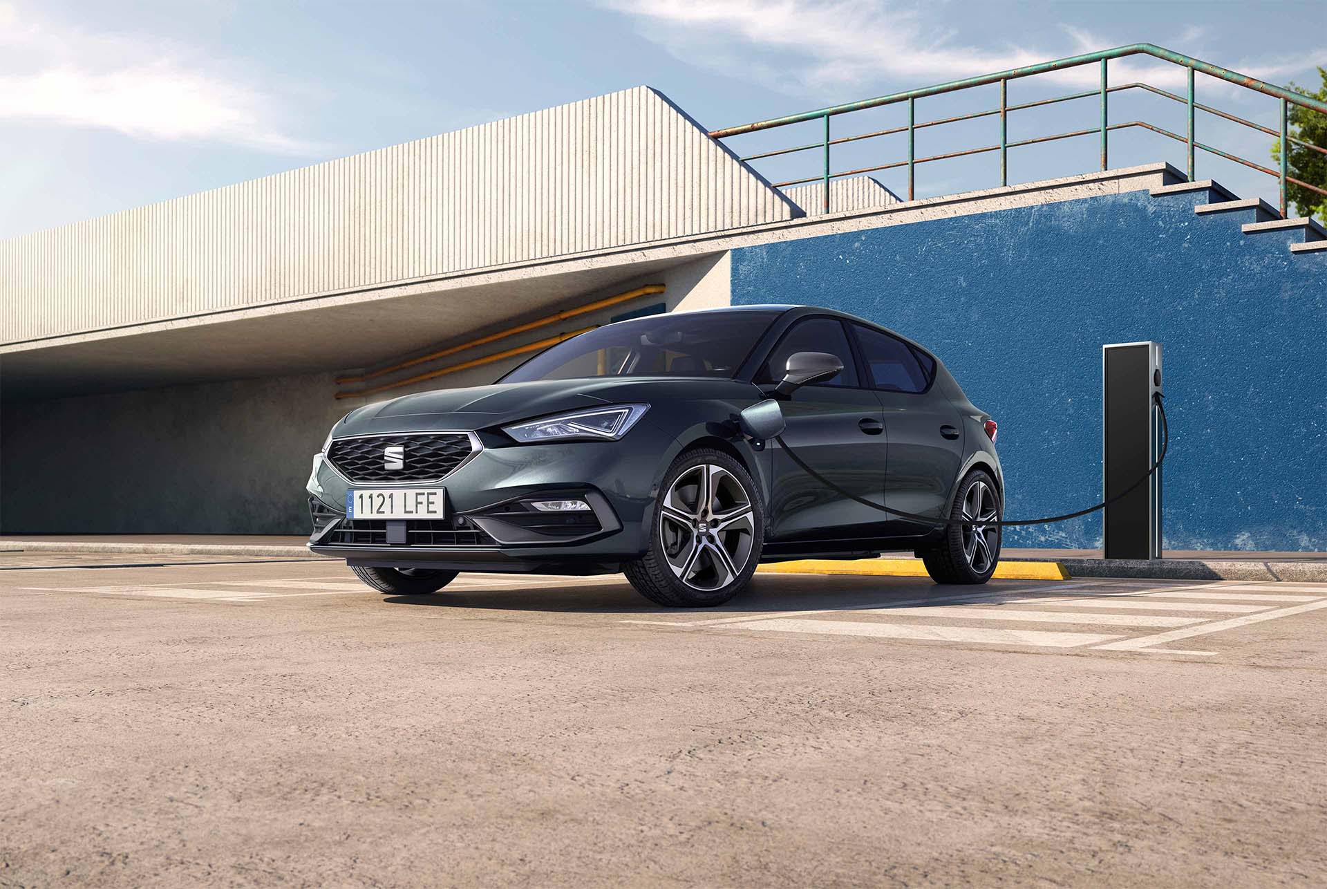 Vue d’une berline compacte sportive SEAT Leon, stationnée en extérieur et en cours de recharge sur une borne noire. Le véhicule se trouve devant un mur bleu imposant avec des escaliers et une passerelle moderne en béton, sous un ciel ensoleillé et partiel