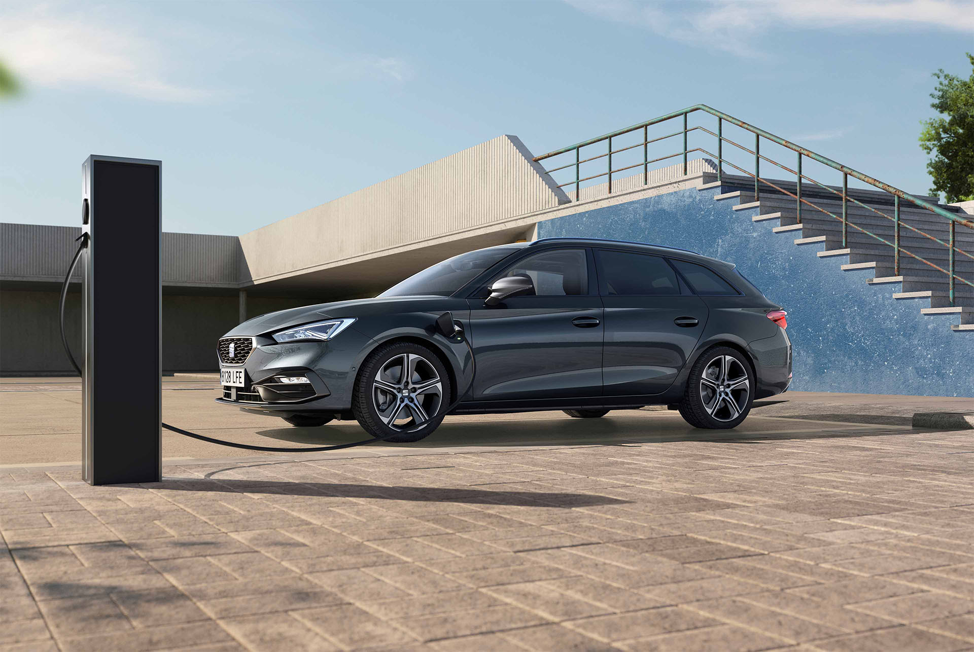 Vue d’une SEAT Leon Sportstourer stationnée sur une zone pavée baignée de soleil et en cours de recharge sur une borne noire. Le break familial hybride rechargeable se trouve dans un environnement extérieur moderne, avec des structures en béton, un escali