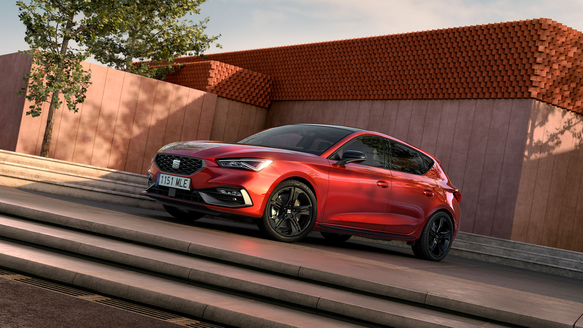 Vue d’une berline compacte sportive SEAT Leon rouge vif, stationnée sur une place en béton baignée de soleil devant un mur en forme de L. Deux palmiers et une architecture moderne minimaliste apparaissent en arrière-plan, sous un ciel bleu dégagé.