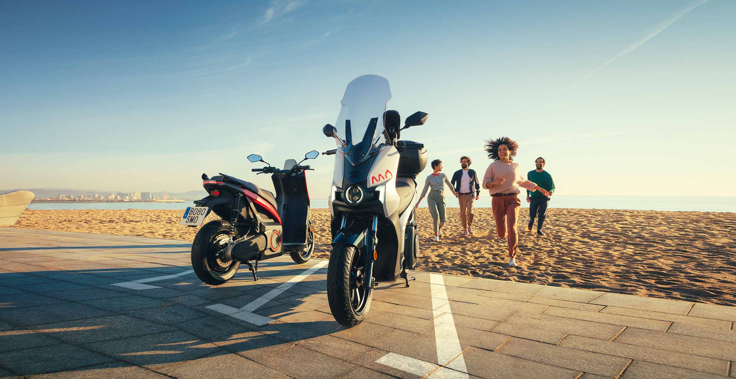 Des amis qui courent vers la MÔ 125 entièrement électrique de SEAT couleur rouge et dark aluminium audacieuse